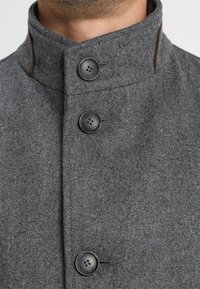 Abrigo de lana gris con un cuello alto, tres botones negros y un acento marrón en contraste con una textura suave.