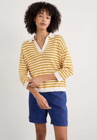 Pull en tricot rayé jaune et blanc avec un col en V et un col blanc, associé à un short en lin bleu avec des manches retroussées.