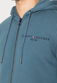 Sudadera con cremallera azul hecha de una tela suave. Presenta una cremallera frontal, cordones y texto de logo bordado en azul marino y rojo.