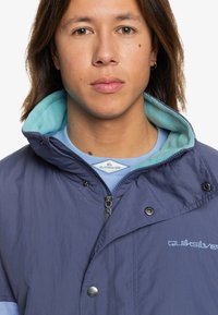 Navy blue jacket with a light blue fleece-lined collar, snap buttons, and embroidered logo on the chest. Features a zippered front.
