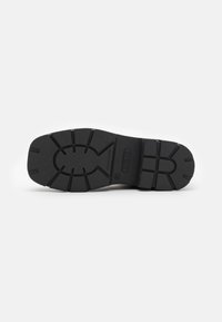 Semelle de chaussure en caoutchouc noir avec un motif de crampons profond, présentant des rainures horizontales et verticales pour une meilleure adhérence. Logo de la marque embossé présent.