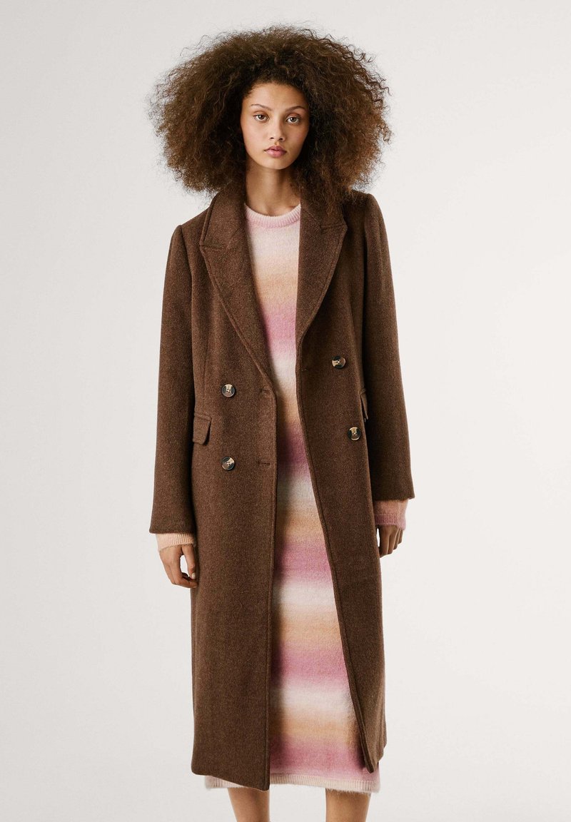 Brown wool double-breasted coat with notched lapels, silver buttons, and side pockets. Worn over a pink and cream striped sweater dress.