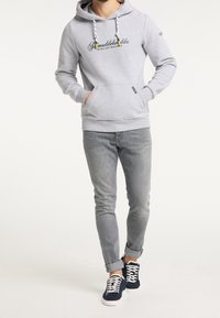 Homme portant un sweat à capuche gris clair avec poche avant, jean gris ajusté et baskets bleu marine avec lacets blancs, debout les mains dans les poches.