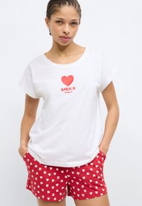 Λευκό βαμβακερό μπλουζάκι με κόκκινη καρδιά και το κείμενο «AMOUR toujours», σε συνδυασμό με κόκκινα σορτς με μικρό λευκό μοτίβο καρδιάς.