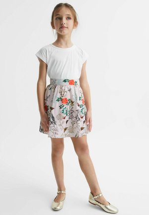 Junges Mädchen steht und trägt ein weißes T-Shirt, einen Rock mit Blumenmuster mit Vögeln und Blumen sowie goldene Mary-Jane-Schuhe auf weißem Hintergrund.