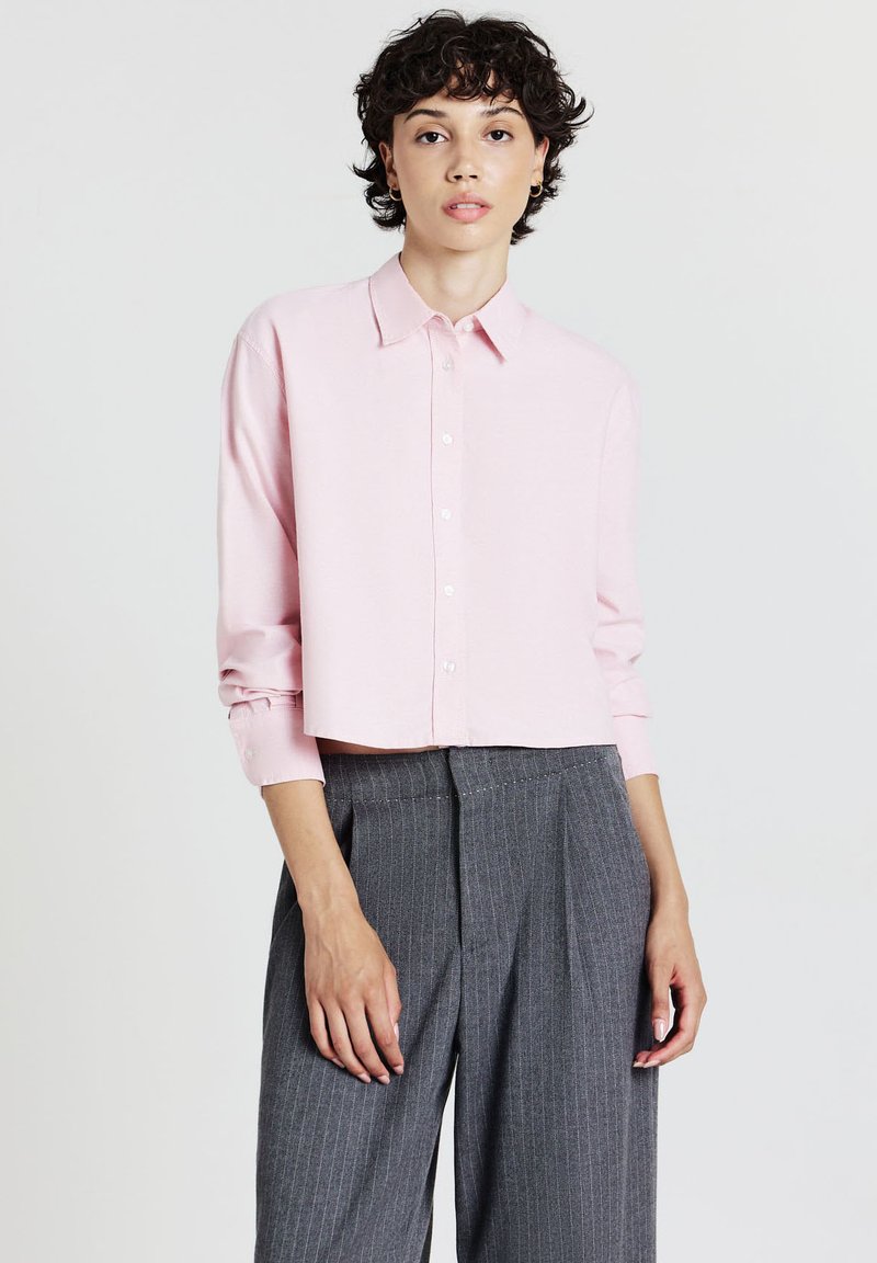 Junge Person in einem hellrosa, kurz geschnittenen Hemd mit Knöpfen und hoch taillierten, grauen, gestreiften, weiten Hosen, die vor einem schlichten Hintergrund steht.