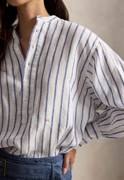 Kvinne iført hvit og blå vertikalt stripete skjorte med delvis knappet krage og blåe bukser med høyt liv og belte.