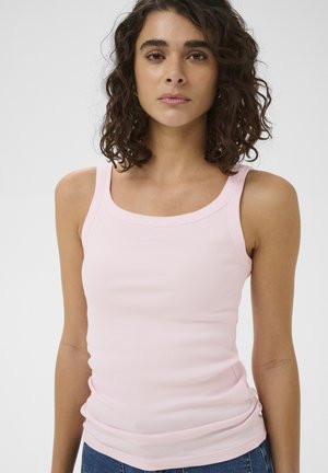 Jeune femme aux cheveux bouclés foncés, portant un débardeur rose clair et un jean bleu, se tenant devant un fond blanc uni.