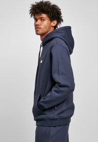 Navy hoodie made of soft cotton blend, featuring a front pocket, drawstring hood, and logo patch on the chest. Smooth texture with ribbed cuffs.