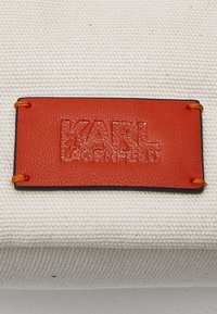 Étiquette rectangulaire en cuir orange avec texte embossé "KARL LAGERFELD", cousue avec un fil orange sur un tissu beige texturé.