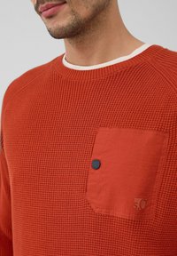 Mann, der einen strukturierten rostorangefarbenen Pullover mit einer festen Brusttasche, Knopfdetails und einem weißen Rundhalsausschnitt-Shirt darunter trägt.