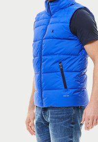 Blå vadderad väst med hög krage, dragkedja framtill och sidoficka. Har horisontell quiltad design och en slät yta.