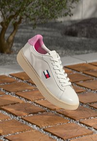Vita lädersneakers med rosa inredning, beige sula, Tommy Hilfiger-logotyp på sidan, placerad ovanför träplattor i kvadrat och små vita stenar.