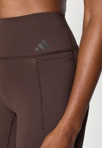 Καφέ αθλητικό κολάν με ψηλό λάστιχο μέσης, επίπεδες ραφές και διακριτική λεπτομέρεια λογότυπου, κατασκευασμένο από λείο, ελαστικό ύφασμα.