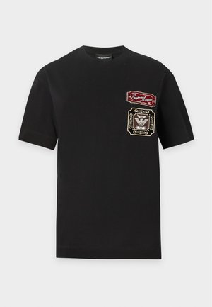 Kobieta stoi, ubrana w czarny t-shirt z nadrukiem, niebieskie jeansy o szerokich nogawkach oraz czarne spiczaste baleriny na prostym jasnym tle.