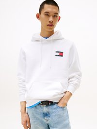 Sudadera blanca con un bolsillo frontal, que presenta un pequeño logo rectangular en rojo, blanco y azul. Lleva puesta sobre una camiseta azul, combinada con unos jeans de denim claro.
