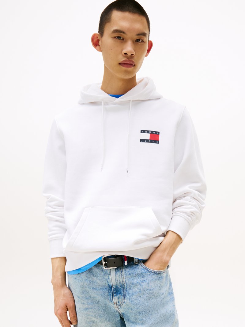 Sudadera blanca con un bolsillo frontal, que presenta un pequeño logo rectangular en rojo, blanco y azul. Lleva puesta sobre una camiseta azul, combinada con unos jeans de denim claro.