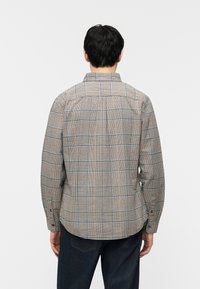 Άνδρας που φοράει μακρυμάνικο μπεζ καρό πουκάμισο και σκούρα τζιν, στέκεται με την πλάτη προς το μέρος μας, μπροστά σε απλό λευκό φόντο.