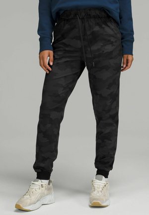 Person iført sorte camouflage joggebukser med elastiske manchetter og hvide sneakers, der står på en grå baggrund.