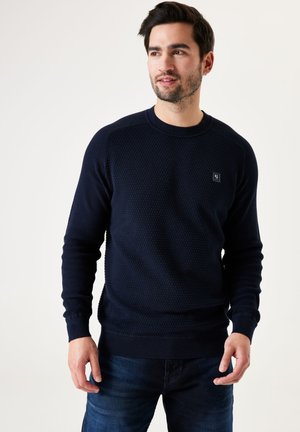 Pull en maille bleu marine avec col rond, bords côtelés aux poignets et à l'ourlet. Présente un petit logo sur la poitrine. Porté avec un jean foncé.