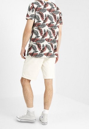Męska koszula z krótkim rękawem z wielokolorowym wzorem liści na jasnym tle, w połączeniu z kremowymi szortami i szarymi trampkami typu high-top.