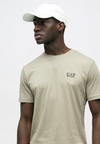 Heledast beežist lühikeste varrukatega t-särk, ümmarguse kaelusega ja väikese musta EA7 logoga rinnal. Kombineeritud valge nokamütsiga.