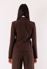 Blazer marrone su misura con silhouette aderente. Presenta maniche lunghe, colletto a revers e una texture liscia. Abbinato a pantaloni coordinati.