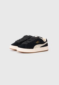 Sneaker in suede nero con suola in gomma crema, caratterizzate da un'accenno decorativo sul Swoosh e spesse stringhe nere. Dettaglio del logo dorato sul lato.