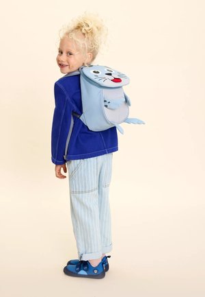 Blue seal-shaped backpack with flippers and facial features. Child wearing a blue jacket and striped pants, paired with blue shoes.