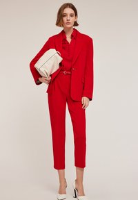 Blazer rouge sur mesure et pantalon assorti, associé à une chemise rouge, portant un sac blanc. Le look est complété par des chaussures à talons blancs.