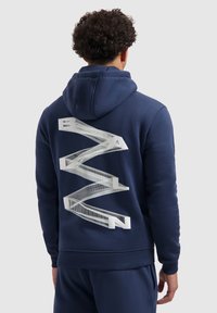 Persoon met krullend haar die een marineblauwe hoodie draagt met een witte 3D zigzagontwerp en letters die "BAWLN" spellen op de rug.
