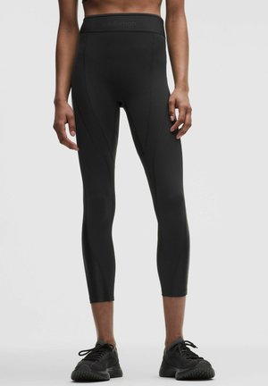 Vooraanzicht van een persoon die zwarte leggings met hoge taille en zwarte sportschoenen draagt, staand tegen een effen lichte achtergrond.