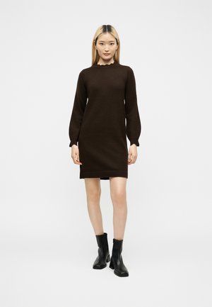 Femme debout portant une robe-pull marron foncé à manches longues avec un col à volants et des bottines noires sur un fond clair uni.