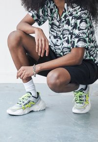 Baskets sportives avec une base blanche, des accents gris et des détails jaune néon. Portées avec un short noir et une chemise à motifs.