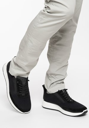 Schwarze Strick-Sneaker mit weißen Sohlen, getragen zu hellbeigen Hosen vor einem schlichten weißen Hintergrund.