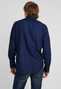 Volwassene met schouderlang bruin haar, gekleed in een lange mouw donkerblauw shirt en vervaagde blauwe jeans, staand met de rug naar de camera tegen een effen achtergrond.