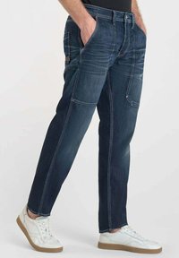 Jeans de mezclilla de color azul oscuro con un corte relajado, que presentan bolsillos frontales, un bolsillo lateral tipo cargo y detalles de costura visibles. Combinados con zapatillas blancas.