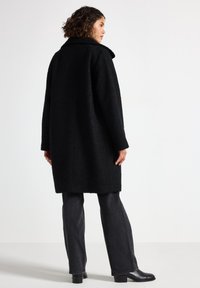 Svart texturerad kappa med bred krage, långa ärmar och rak siluett. Kombinerad med mörkgrå utsvängda jeans och ankelstövlar.