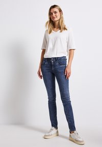 Jean skinny bleu avec un lavage moyen, associé à un t-shirt blanc à col en V. Des baskets blanches décontractées complètent le look, soulignant le confort.