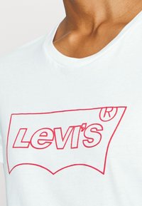 Vit bomull t-shirt med rund halsringning och en framträdande röd Levi's-logotyp i en klassisk design över bröstet. Slät textur.
