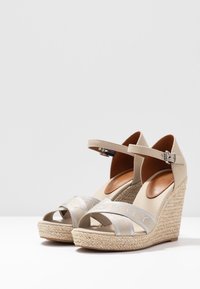 Beige wedge sandalen met een geweven jute zool. Voorzien van metallic zilveren bandjes en een verstelbare enkelgesp. Glad oppervlak en open teen ontwerp.