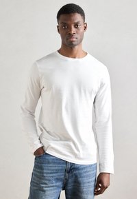 Hvit langermet t-skjorte laget av mykt stoff, med rund hals og en tettsittende design. Kombinert med lys blå denimjeans.