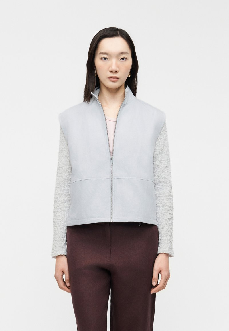 Femme aux cheveux bruns et raides porte une veste sans manches gris clair zippée par-dessus un haut texturé à manches longues et un pantalon marron foncé, debout droit.