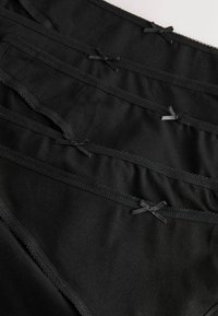 Crni pamučni donji rublje glatke teksture, s ukrasnim valovitim rubom i malim mašnicama na pojasu. Više pari složenih u hrpu.