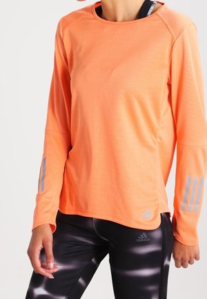 Osoba nosząca pomarańczowy sportowy top z długim rękawem z odblaskowymi paskami oraz czarne legginsy Adidas z białymi wzorami.