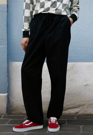 Personne portant un pantalon noir, une chemise à carreaux à manches longues et des baskets rouges, debout contre un mur bleu et blanc.