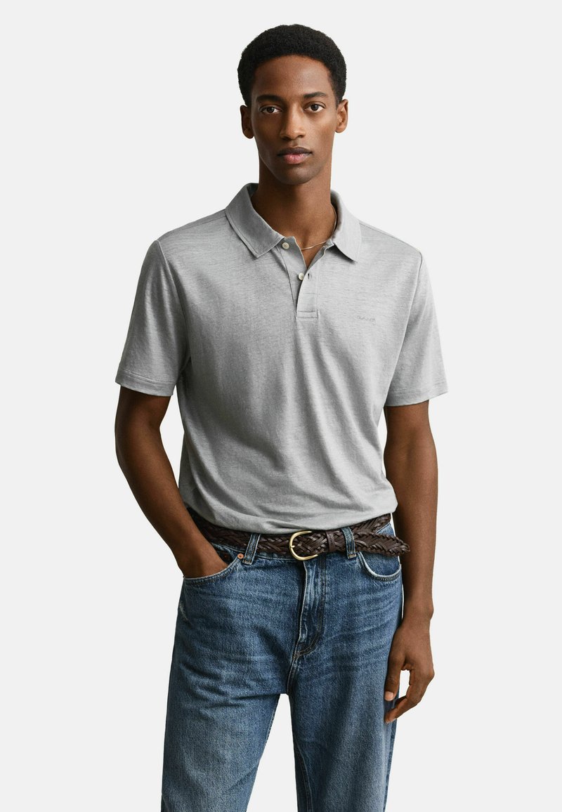 Junger Mann trägt ein hellgraues Poloshirt, das in blaue Jeans mit einem geflochtenen braunen Gürtel gesteckt ist, steht mit einer Hand in der Tasche.