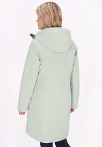 Pardessus impermeabile verde chiaro con cappuccio, cuciture sulle spalle e vita con cordoncino. Caratterizzato da un design di lunghezza media e una texture liscia.