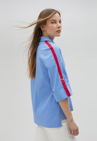 Camicia oversized azzurro chiaro con maniche arrotolate, caratterizzata da una striscia rossa lungo il braccio e dettagli in metallo argentato. Tessuto morbido, design casual.