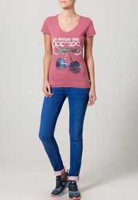 Rosa V-ringad t-shirt med vit text och paljettdesign av körsbär. Buren med blå jeans och svarta sportskor med orange detaljer.